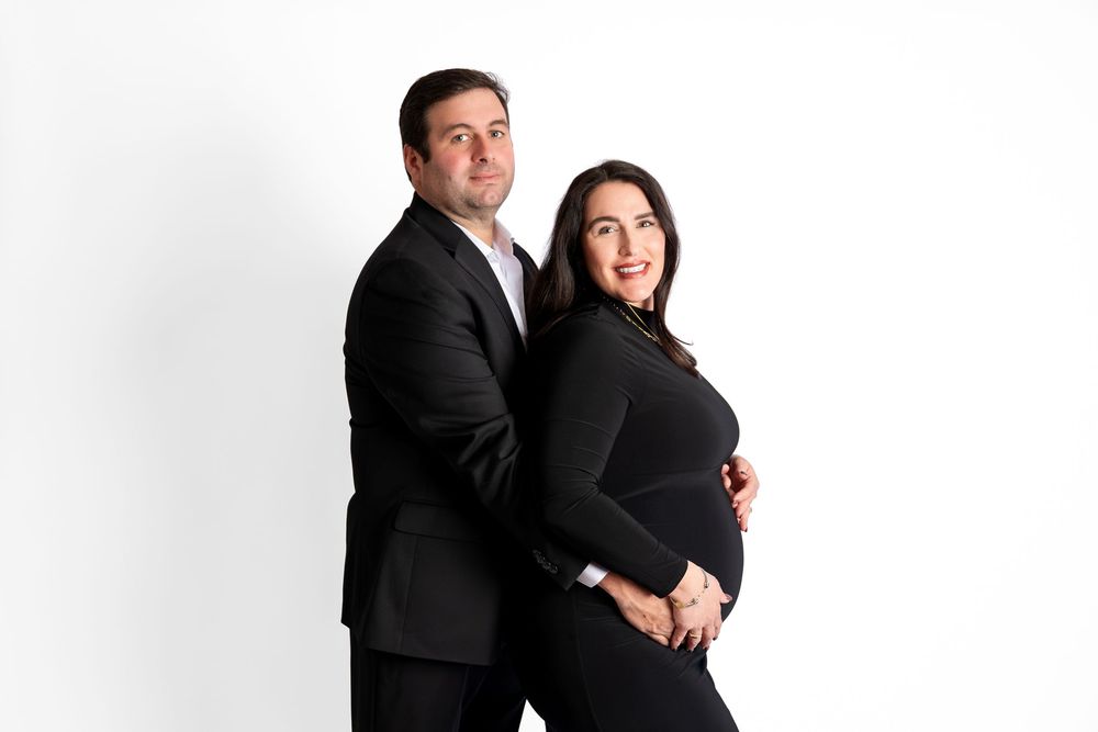 Couple in black outfits posing against white background, pregnant woman cradling baby bump while man stands back-to-back with her.