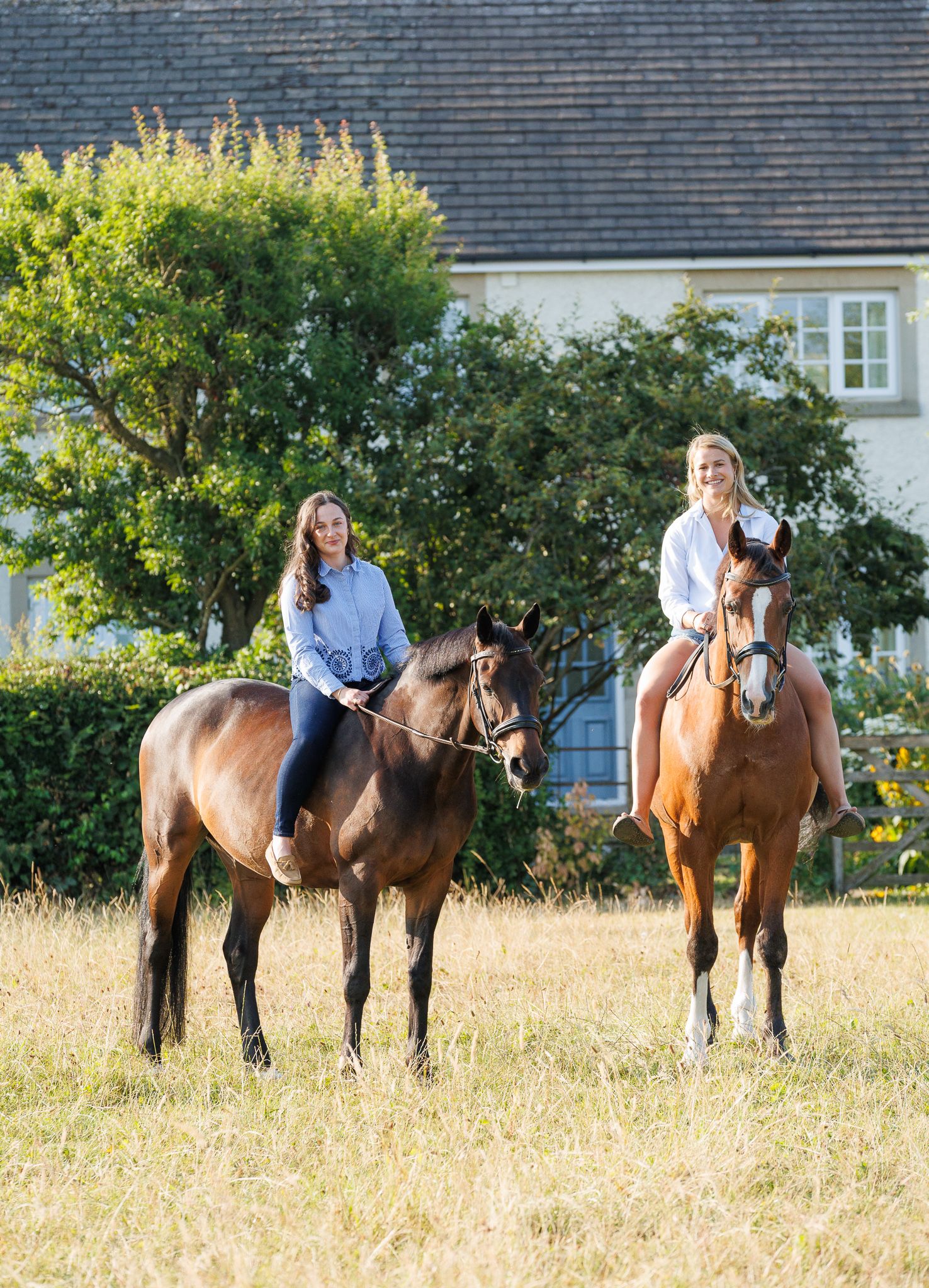 Woman walking two chestnut horses down a sunlit gravel path lined with green trees at a horse farm.
