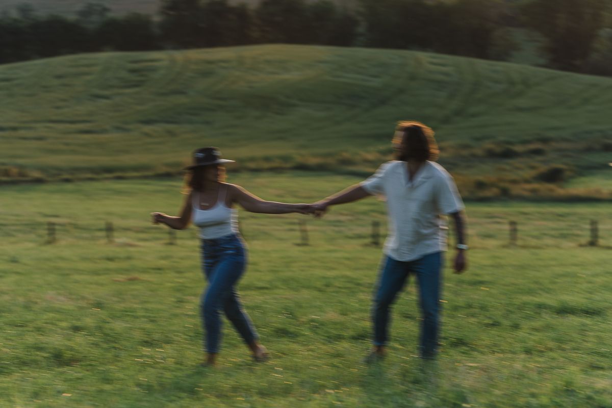 Couple in jeans holds hands and walks across a green grassy field with rolling hills and trees in the background.