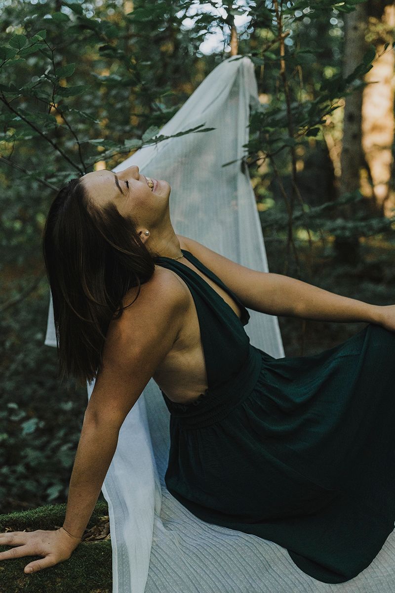 Woman in a dark green dress tilts her head back on a forest log with white fabric flowing behind her.