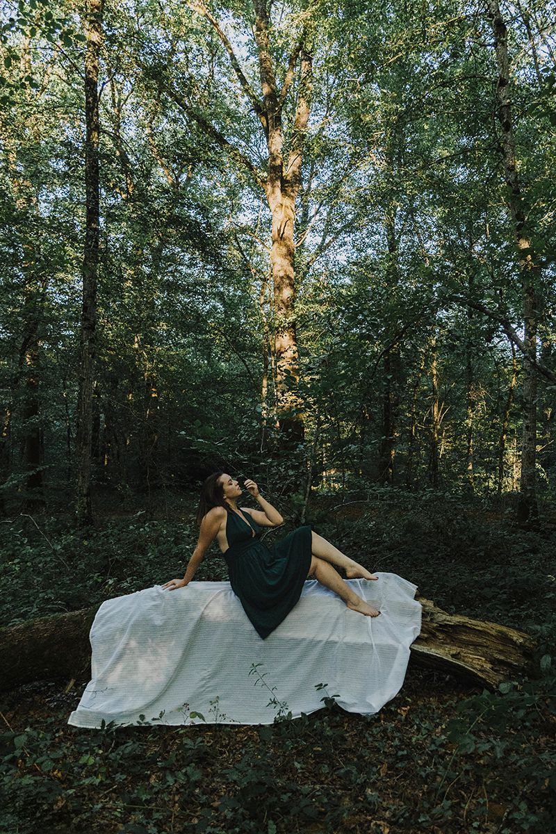Woman in a dark dress poses on a white cloth over a fallen log surrounded by tall green forest trees.
