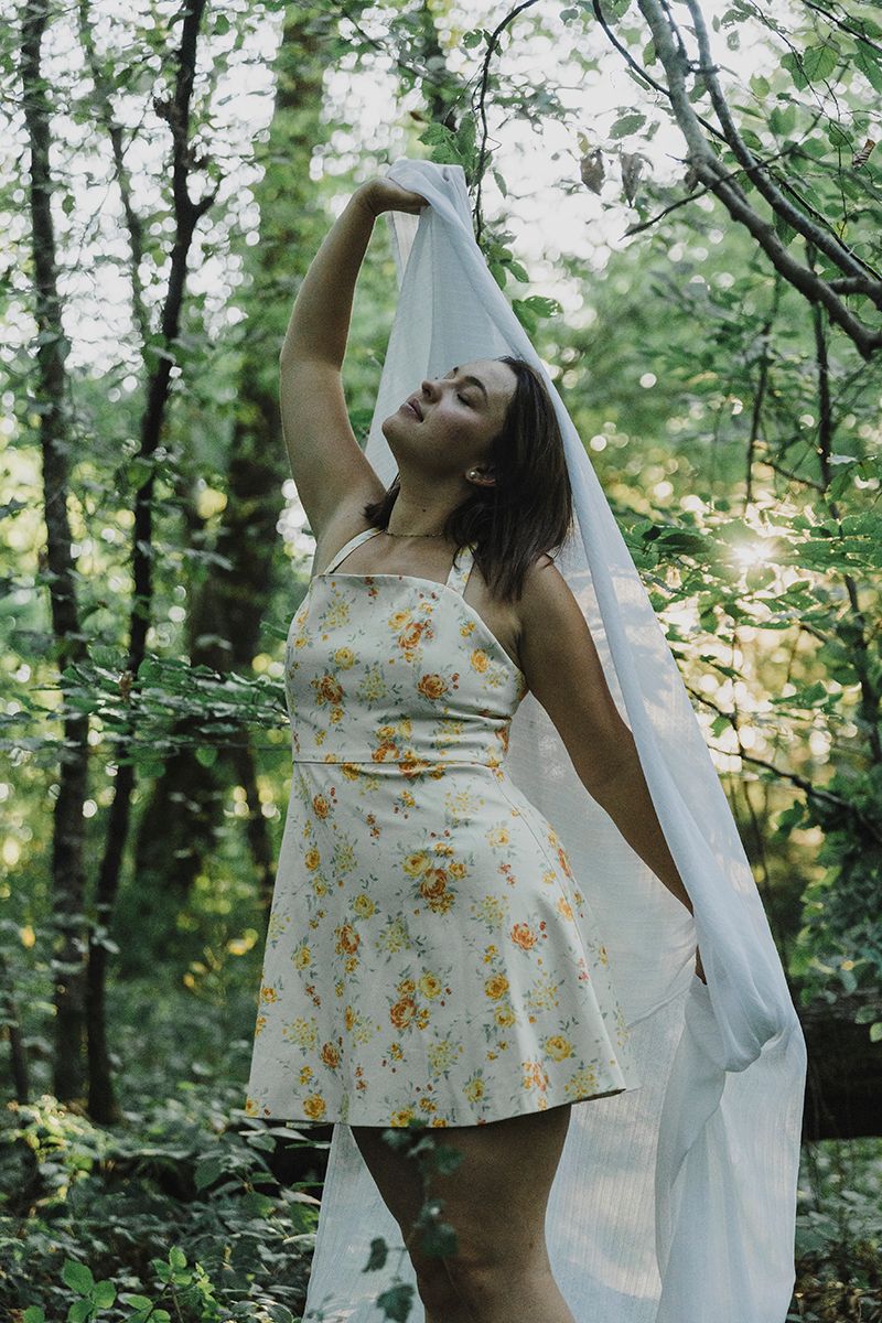 Woman in a yellow floral sundress raises a sheer white veil among sunlit green trees in the forest.