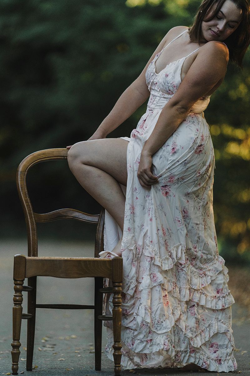 Woman in a ruffled floral dress rests one foot on a wooden chair outdoors with a blurred green background.