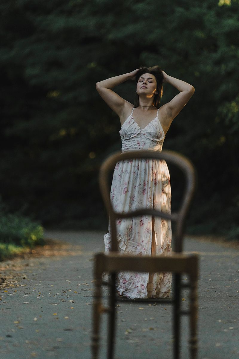 Woman in a long floral dress stands on a forest road with her arms raised, an empty wooden chair in the foreground.