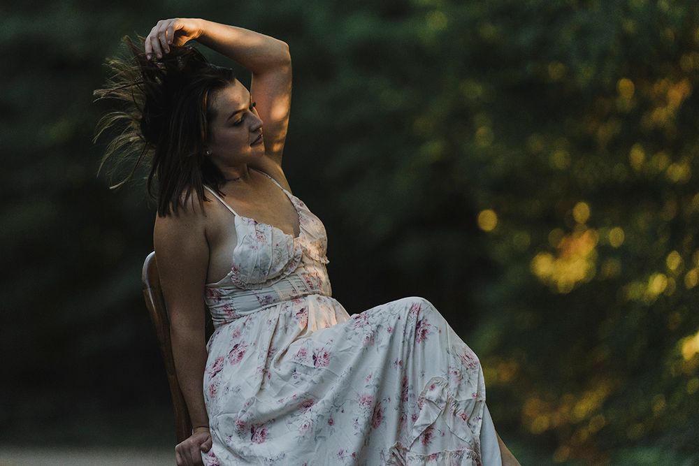 Woman in a floral sundress sits on a wooden chair outdoors, running her hand through her hair in warm golden forest light.