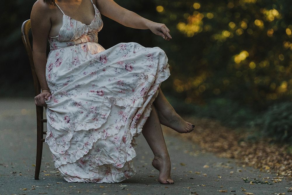 Woman in a flowing floral dress sits on a wooden chair on a path, one bare foot lifted, with golden foliage glowing behind.