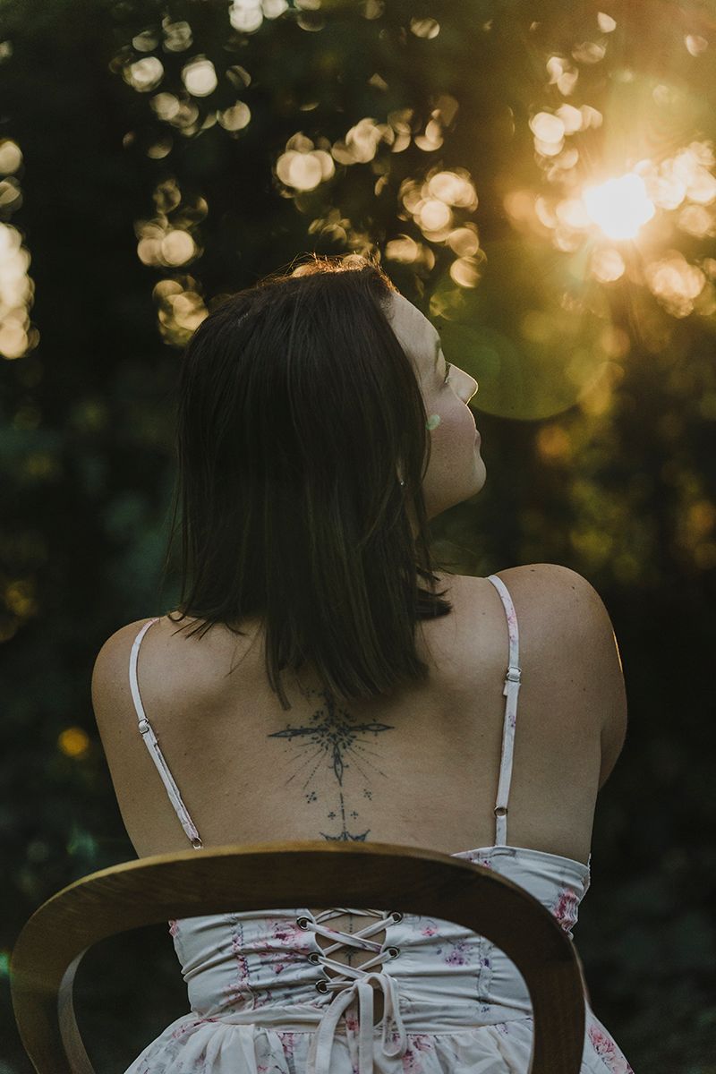 Back view of a woman with short dark hair and a tattoo on her back, sitting on a chair against sunlit forest bokeh.