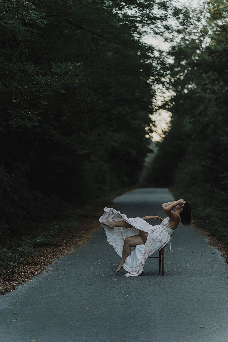 Woman in a white dress tips back on a wooden chair in the middle of a tree-lined road as her skirt billows upward.