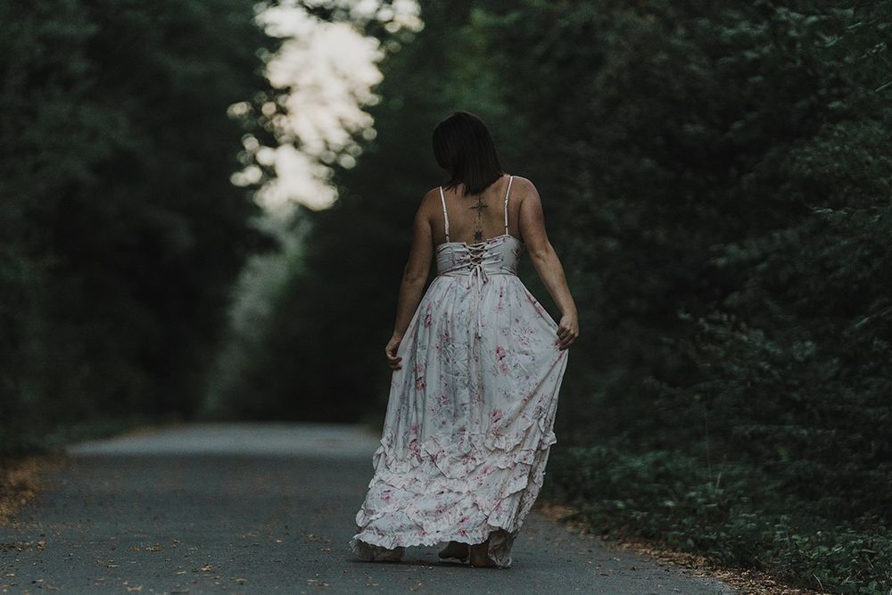 Woman in a flowing floral dress walks away along a quiet tree-lined road, holding her skirt, in soft dusk light.