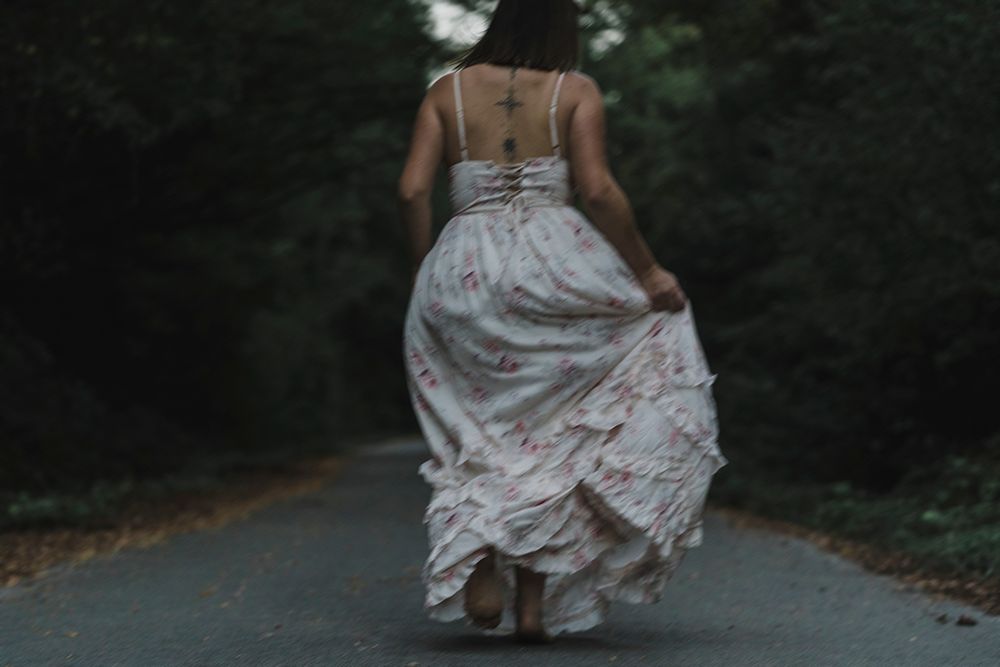 Barefoot woman in a long floral dress runs away down a dark forest road, her skirt swirling behind her.