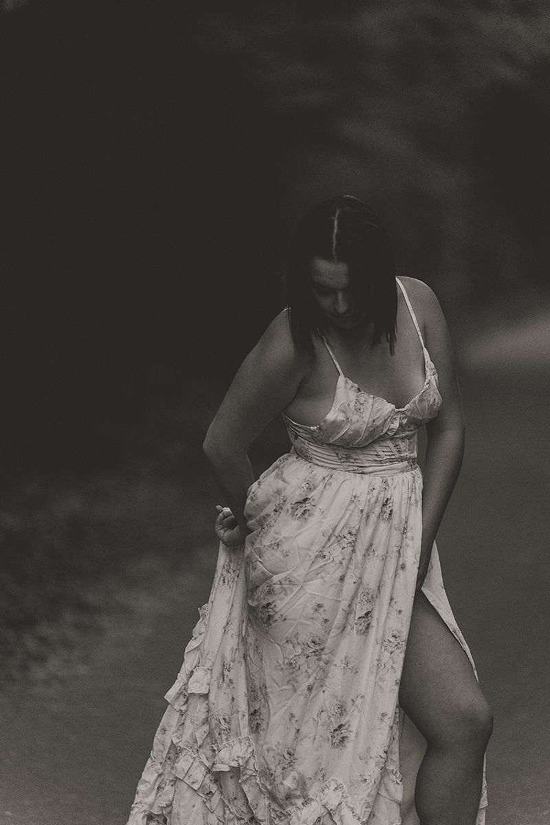 Black and white portrait of a woman in a ruffled floral dress with a thigh-high slit, gathering her skirt and looking down.