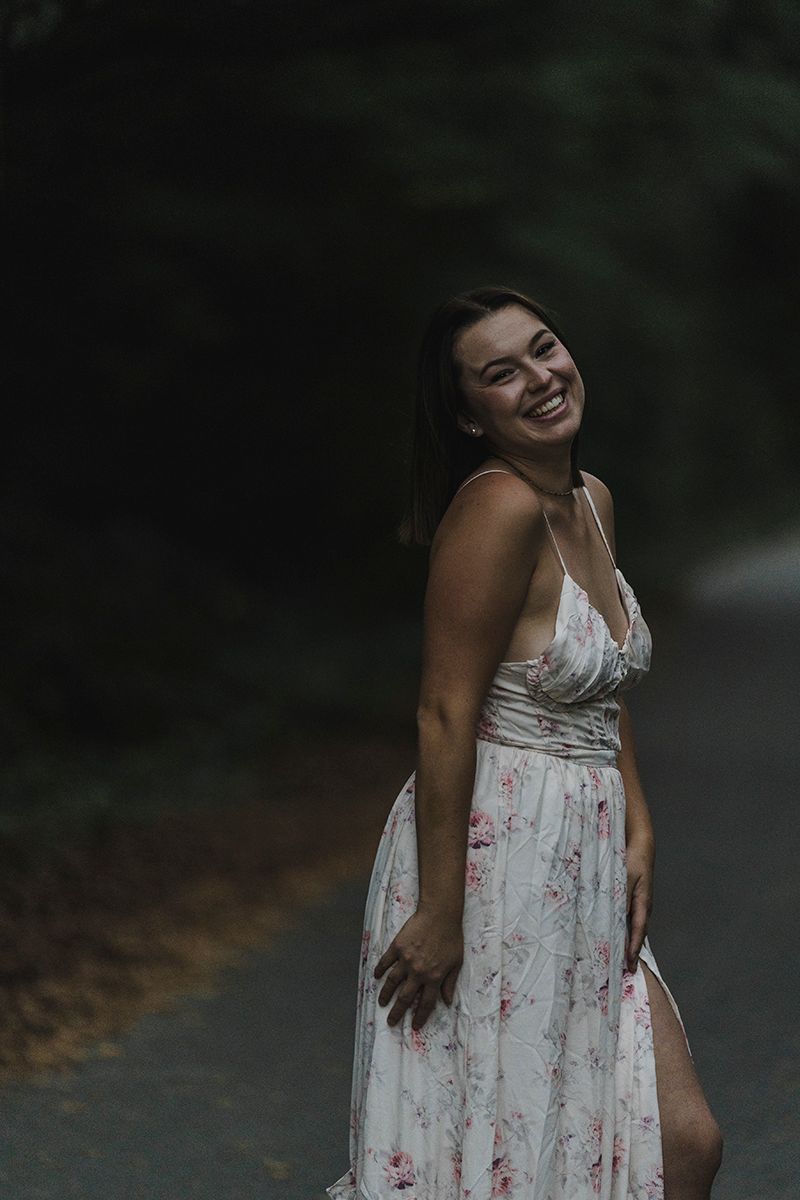 Smiling woman in a white floral sundress with a leg slit stands on a shaded forest road, laughing with her head tilted.