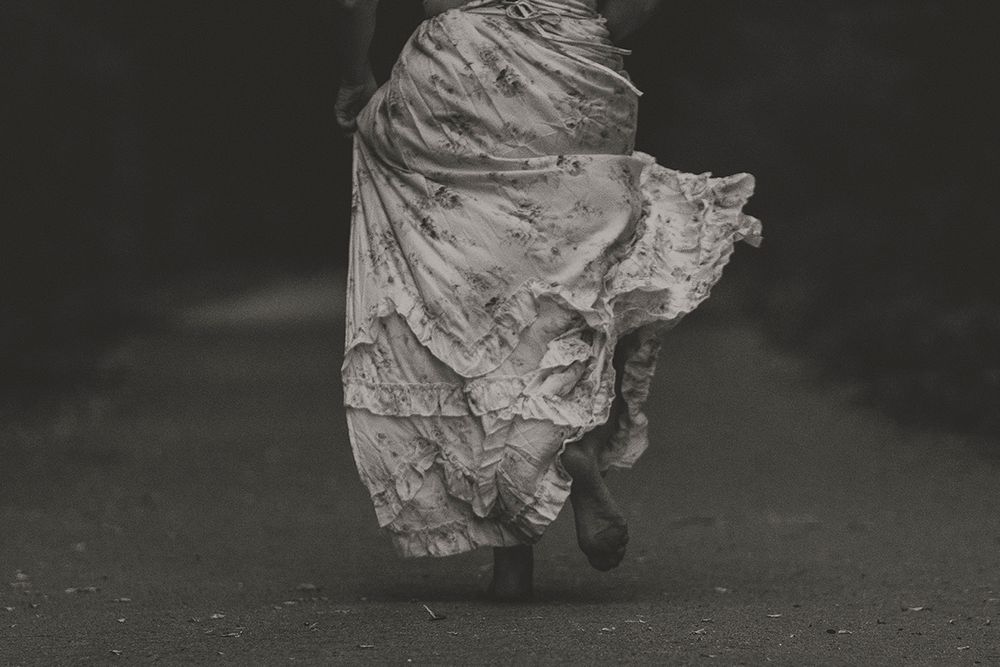 Black and white photo of a woman's bare feet and tattered floral skirt in motion as she runs along a dark road.