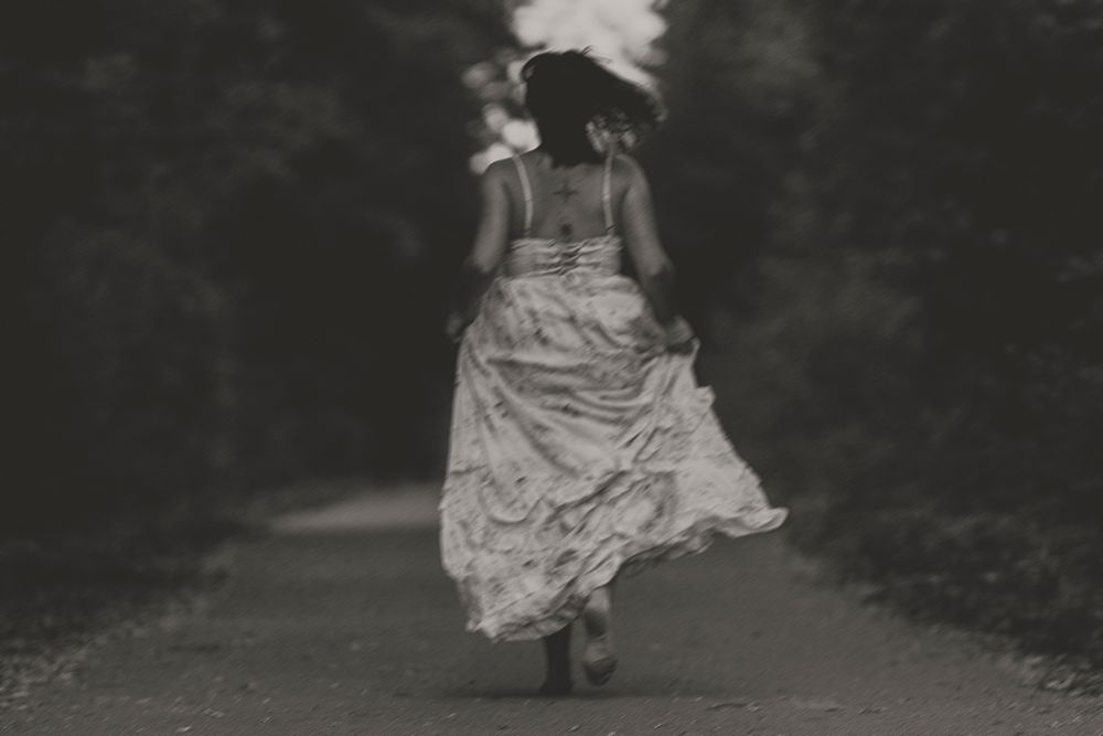 Black and white photo of a woman in a flowing long dress running barefoot down a tree-lined path away from the camera.