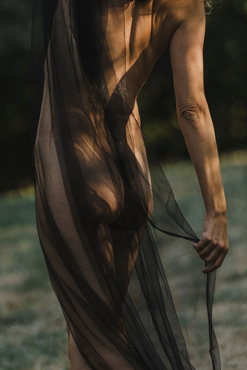 Artistic back view of a figure draped in sheer black tulle fabric, holding the cloth's edge in soft dappled sunlight outdoors.