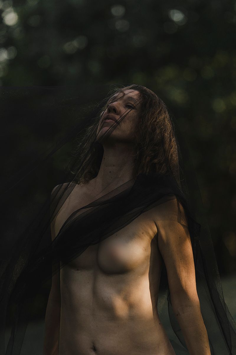 Moody portrait of a woman with long hair in a sheer black veil, face tilted upward in dim, dappled forest light.