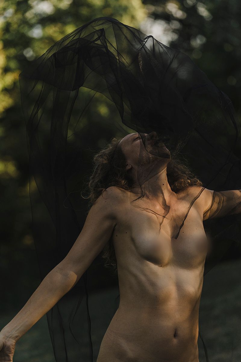 Woman with curly hair tosses back her head as sheer black fabric billows around her in a sunlit green woodland.