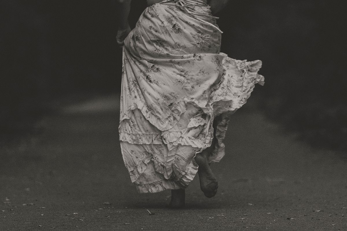 Black and white photo of a person in a tattered, flowing vintage skirt running barefoot along a dark country road.