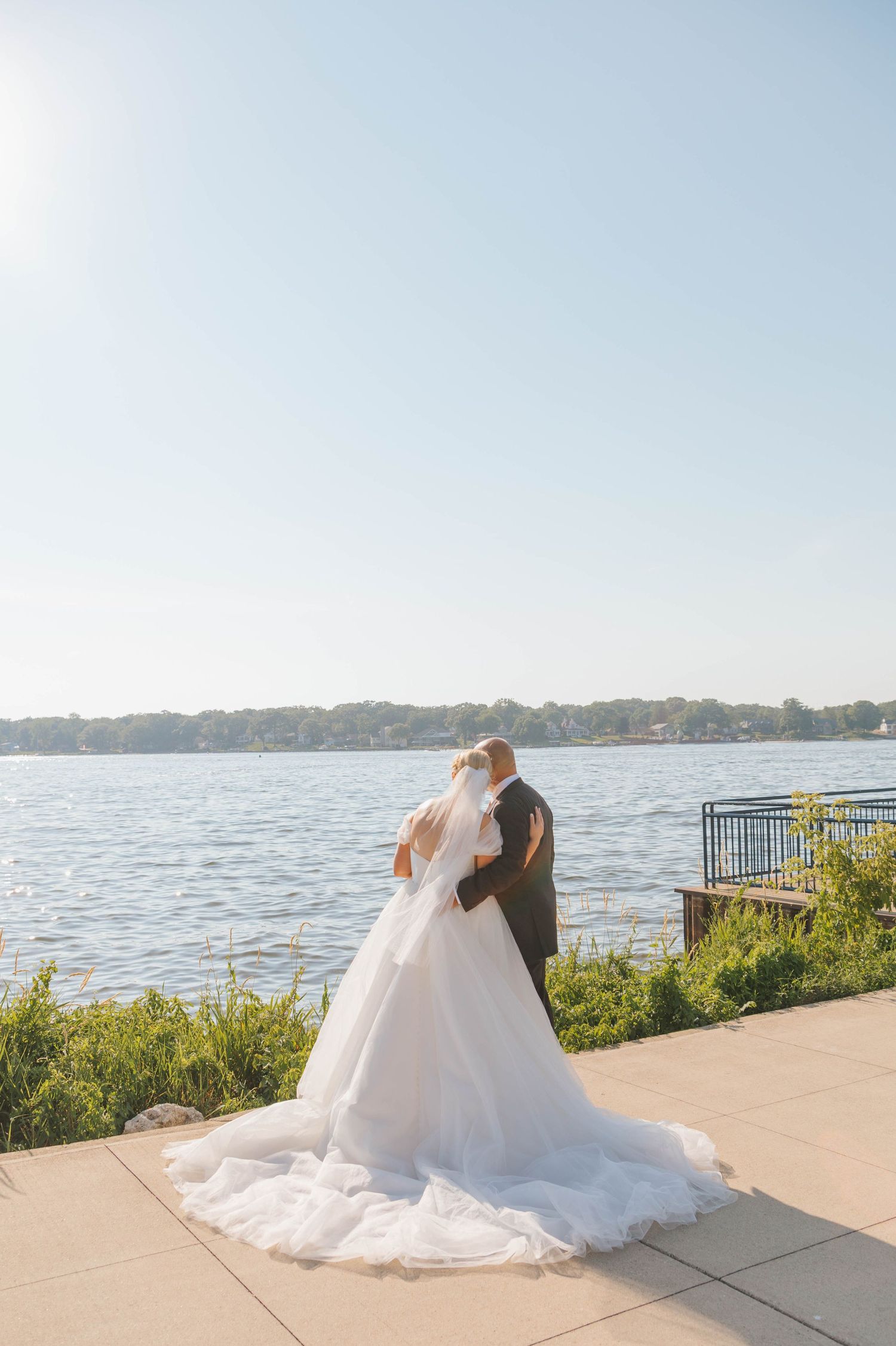 Bride and groom embrace facing a serene lake on their wedding day, bride wearing a flowing white gown with long train.