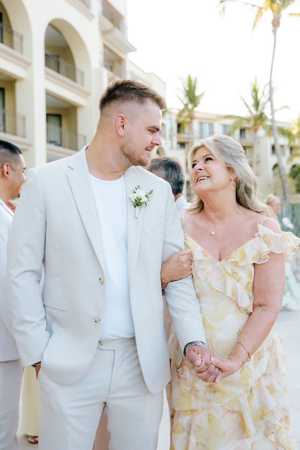 Smiling groom in a light suit with a floral boutonniere walks arm in arm with a woman in a yellow floral dress at a tropical resort wedding.