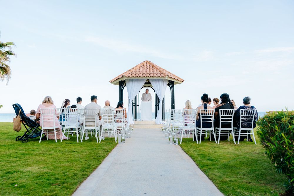 Wedding guests seated in white chairs on a lawn facing a tropical beachfront gazebo with draped white curtains and a terracotta roof.