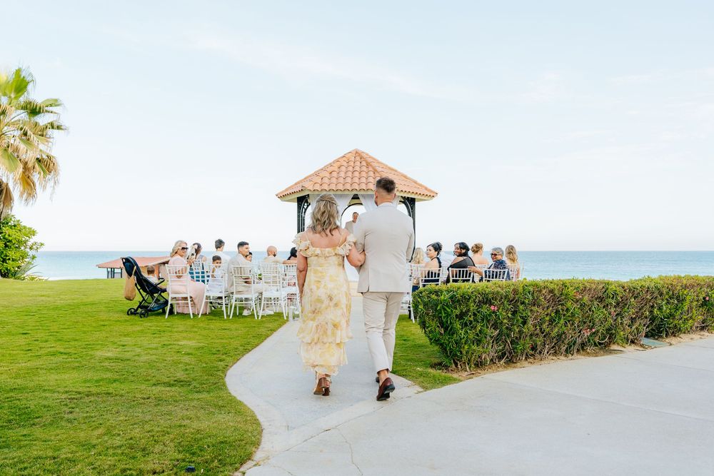 Couple walks arm in arm down a paved path toward guests seated by an oceanfront gazebo during a seaside wedding ceremony.