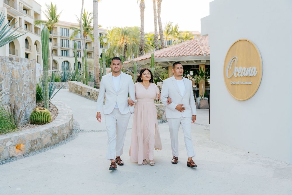 Bride in a blush pink gown walks arm in arm with two men in white suits along a palm-lined path at a sunny desert resort wedding venue.