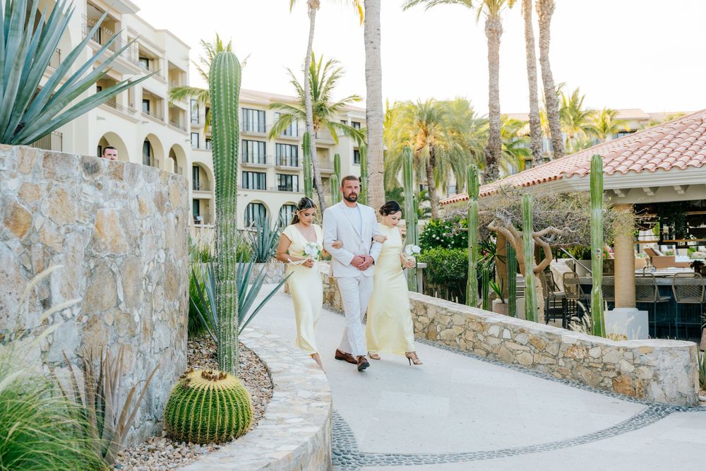 Bride and groom with a woman in flowing cream gowns walk a stone path past cacti and palm trees at a tropical resort.