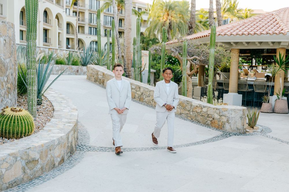 Two young men in light gray suits walk along a stone pathway at a tropical resort, lined with cacti, palm trees, and a covered terrace.