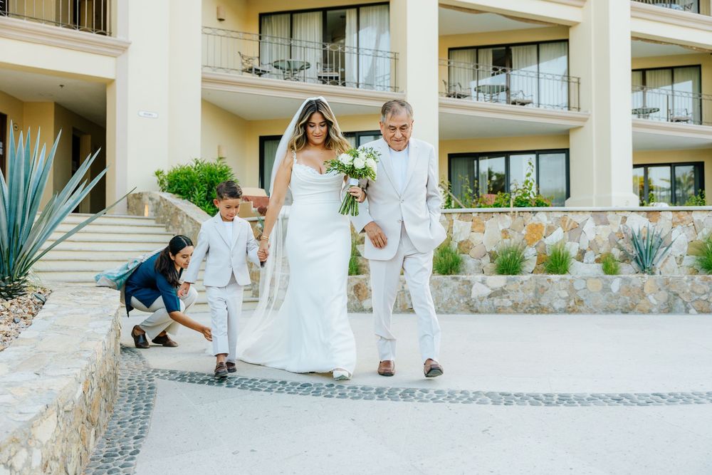 Bride in a white gown and veil carrying a bouquet walks with an older man in a light suit past a young boy at a resort wedding.