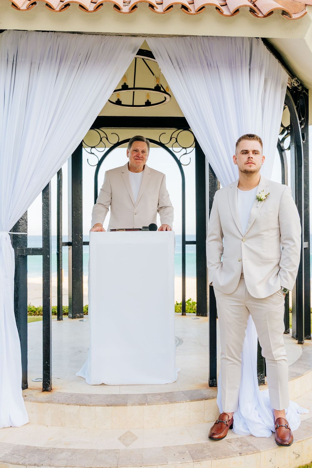 Groom in a cream suit with boutonniere stands beside an officiant at a podium in a white-draped beachfront wedding gazebo.