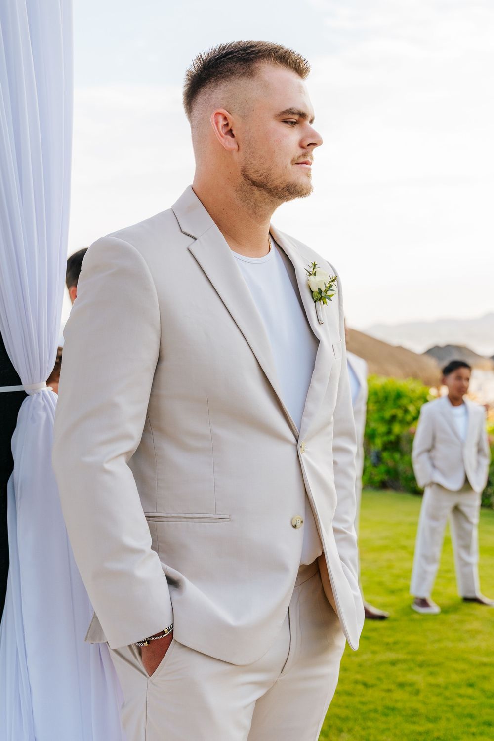 Groom in a light beige suit with a floral boutonniere stands with hands in pockets at an outdoor wedding ceremony on a sunny lawn.