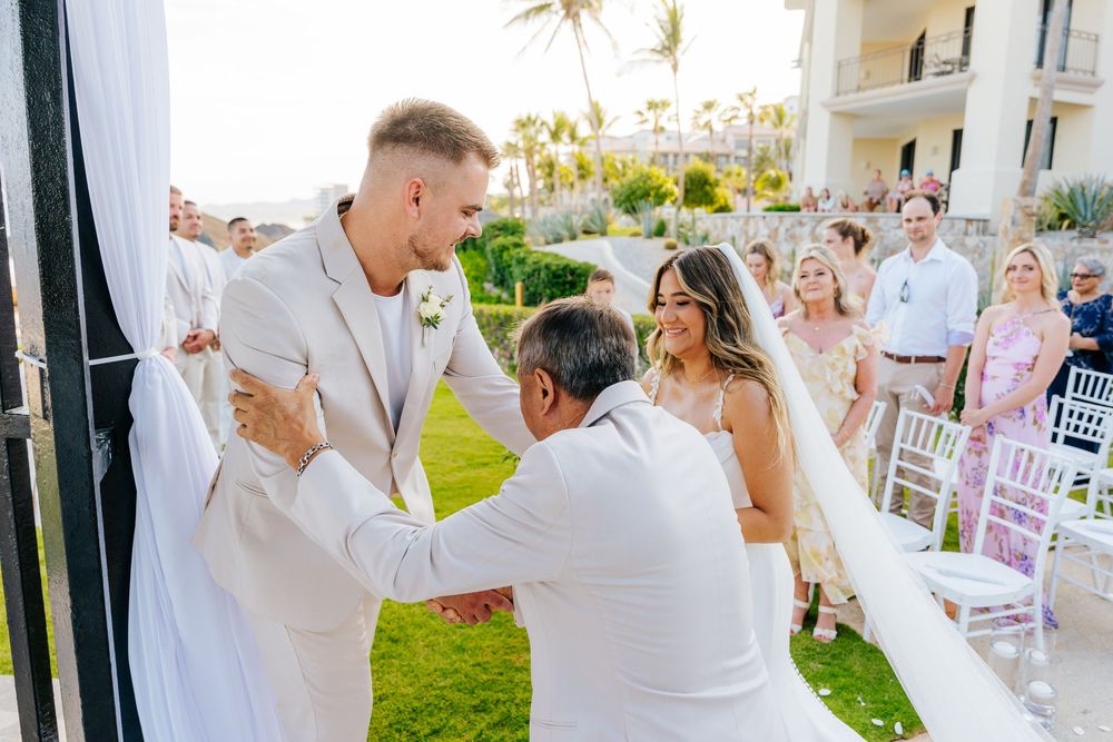 Groom in a light suit shakes hands with an older man in white as the bride smiles beside them at a tropical beachside wedding ceremony.