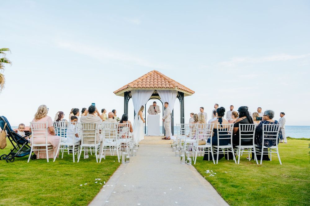 Outdoor oceanfront wedding ceremony with guests seated on white chairs along an aisle leading to a tiled-roof gazebo on green grass.