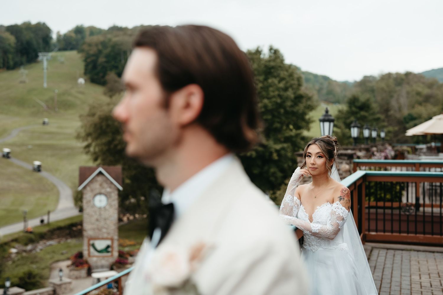 Bride in lace gown gazes at groom in white suit during outdoor wedding at scenic mountain resort.