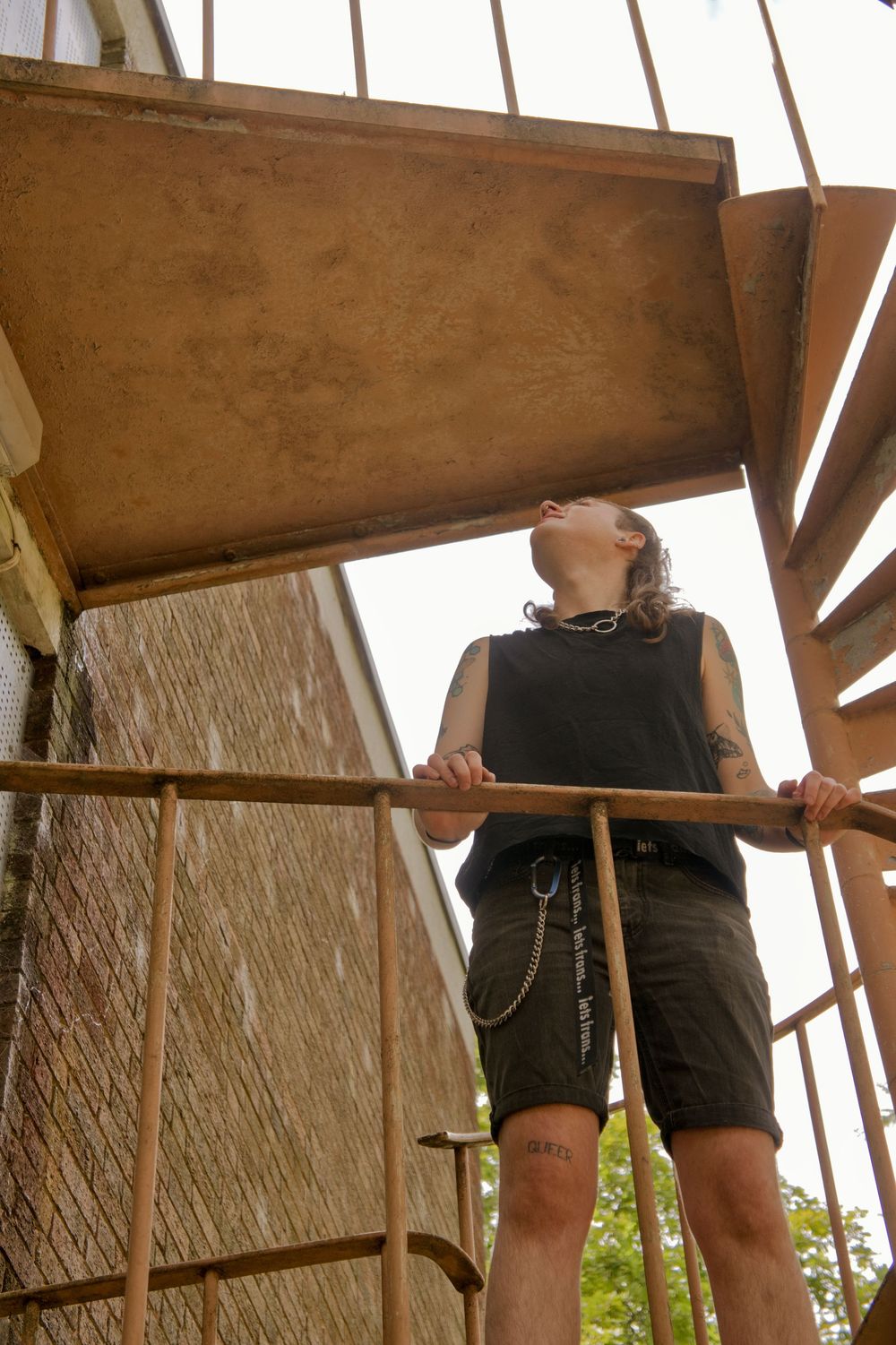 Tattooed person in black sleeveless shirt looking up at rusty spiral staircase on brick building exterior.