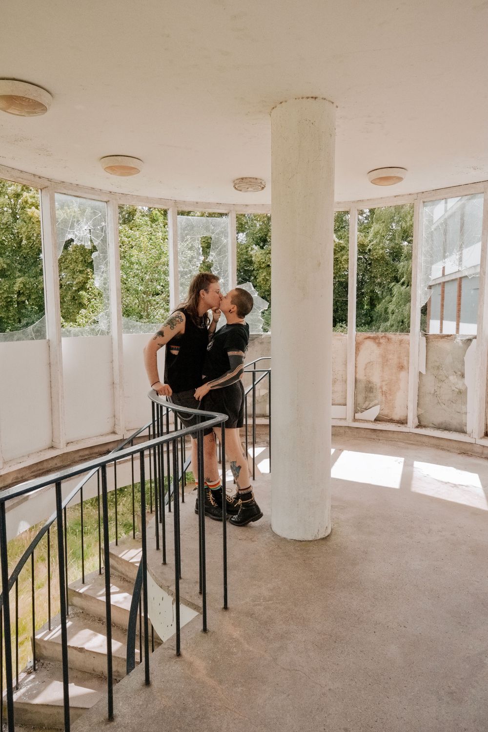 Two people sharing a kiss on a staircase landing in an abandoned building with large windows.