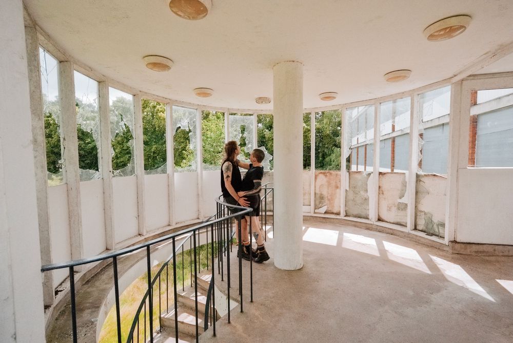 Couple embracing on a curved white balcony with tall columns and large windows in an elegant abandoned building.