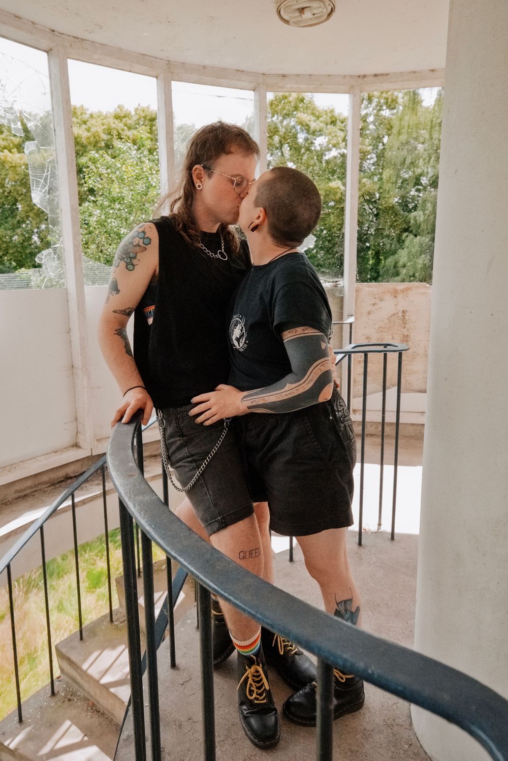 Two tattooed people sharing a kiss on a spiral staircase in a bright, windowed space.