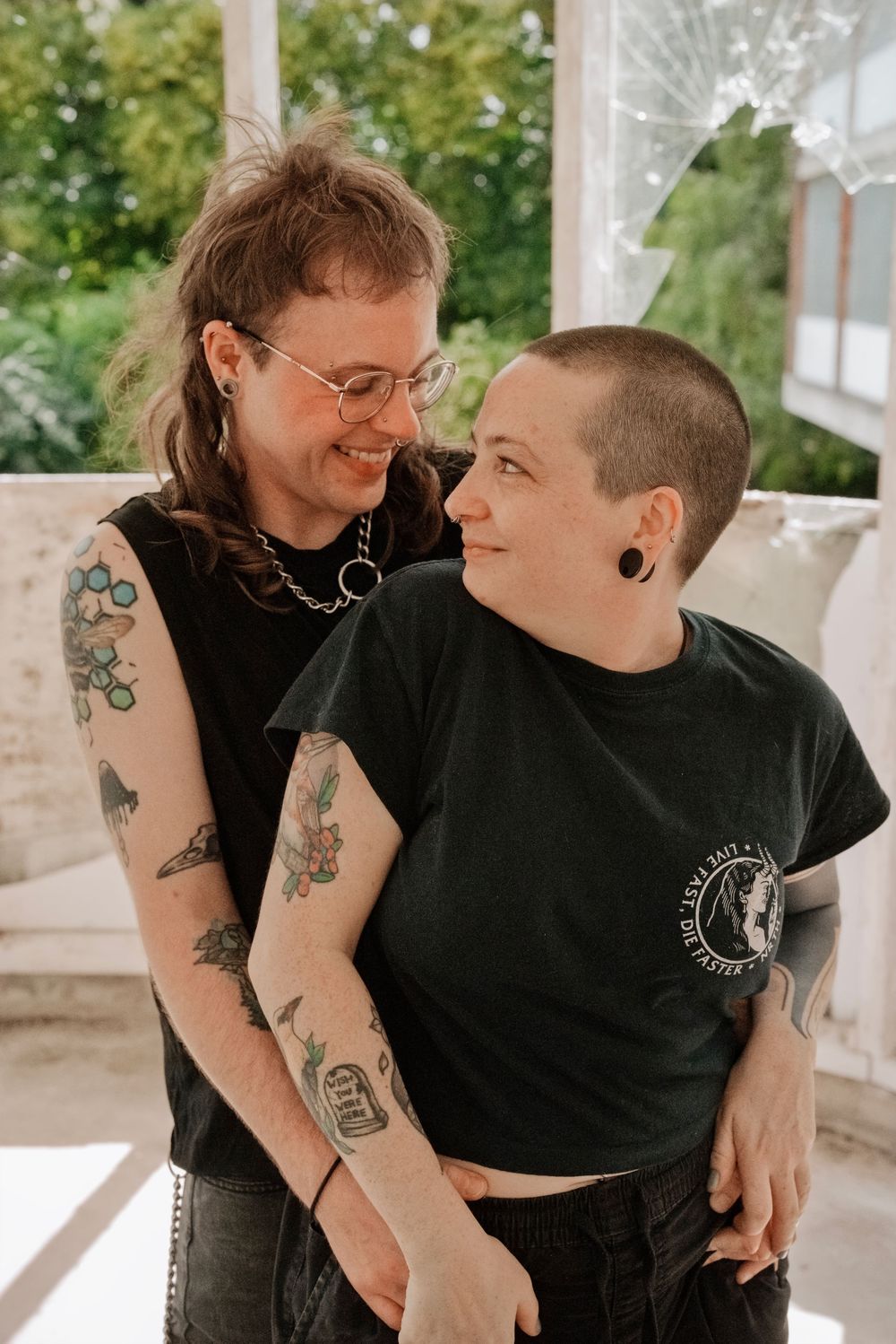 Two tattooed people in black shirts smiling and embracing indoors near bright windows with greenery.