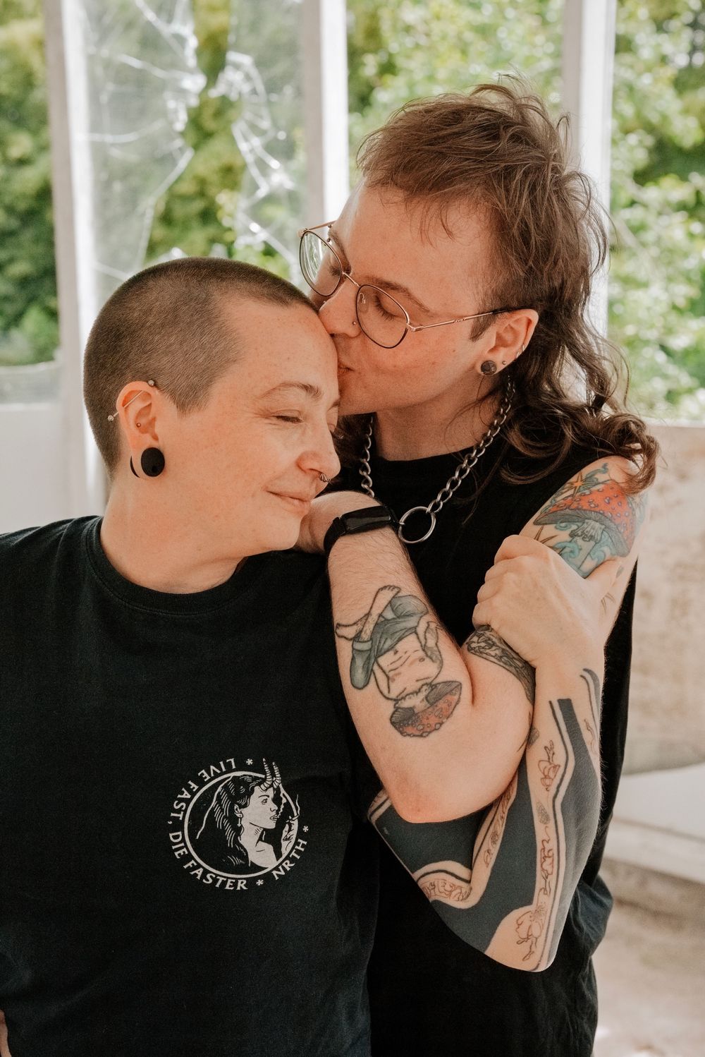 Two tattooed partners share a tender kiss, one with a shaved head and gauge earrings, both wearing black shirts.