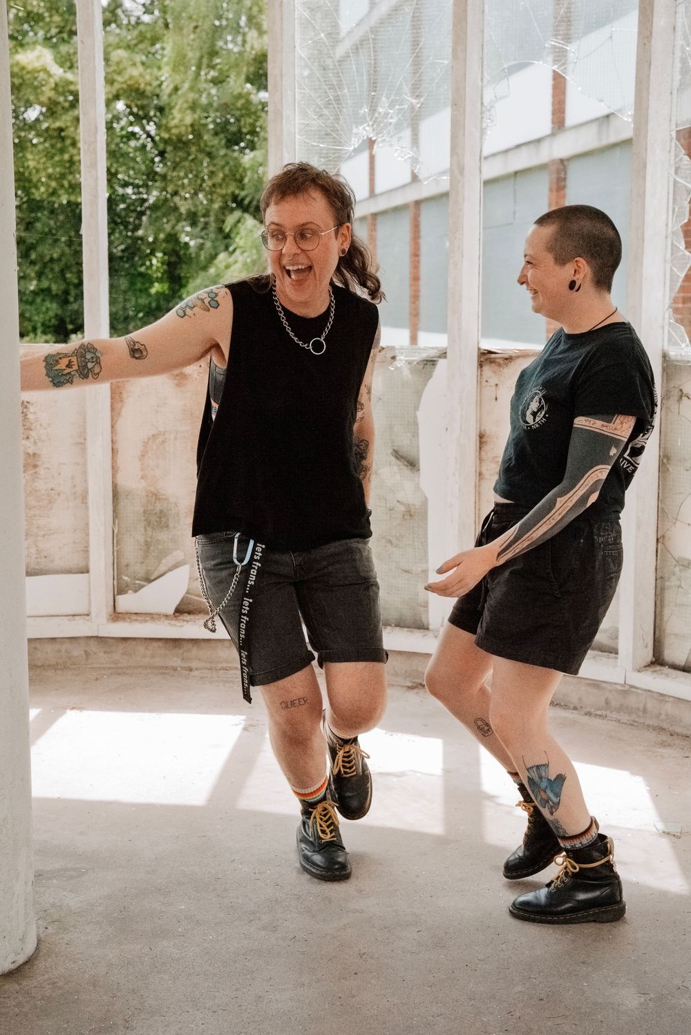 Two tattooed friends laughing and dancing together outdoors in black casual outfits and boots.