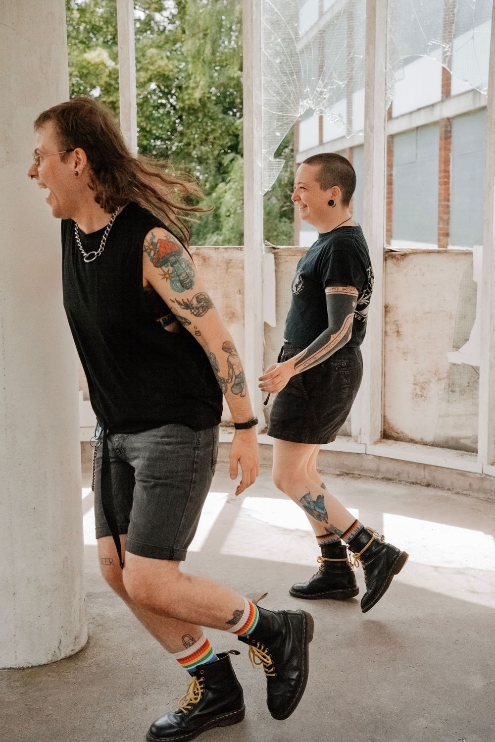 Two tattooed people in black outfits and Doc Martens walk through a sunlit building with large windows.