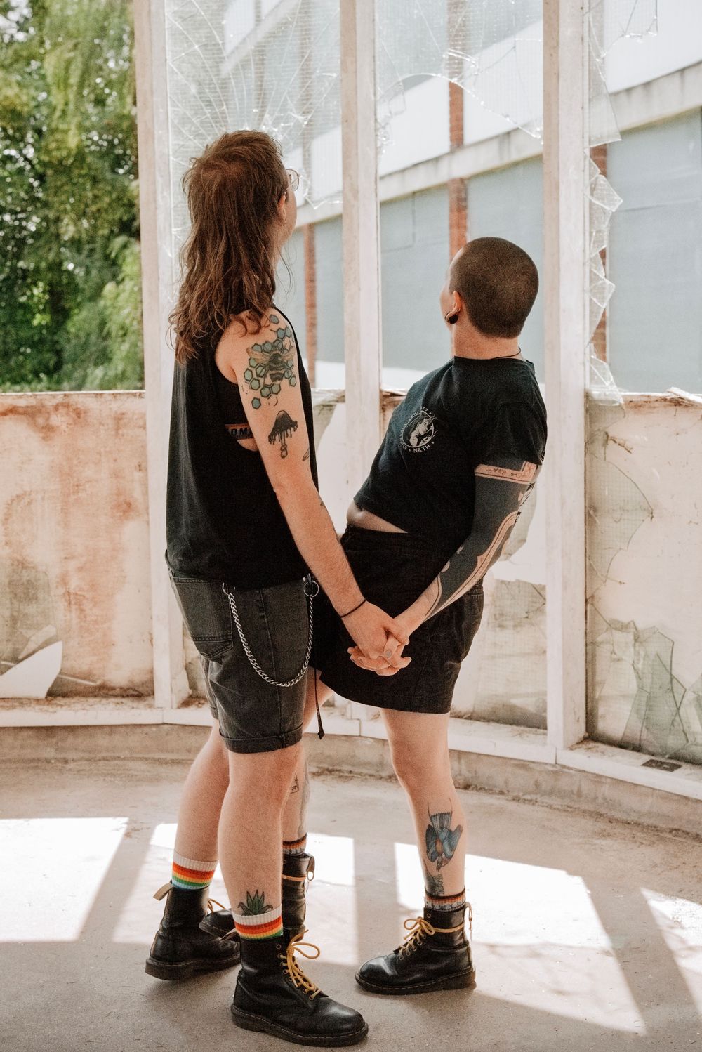 Two tattooed people holding hands in black outfits and Doc Martens boots, standing in a sunlit abandoned building.