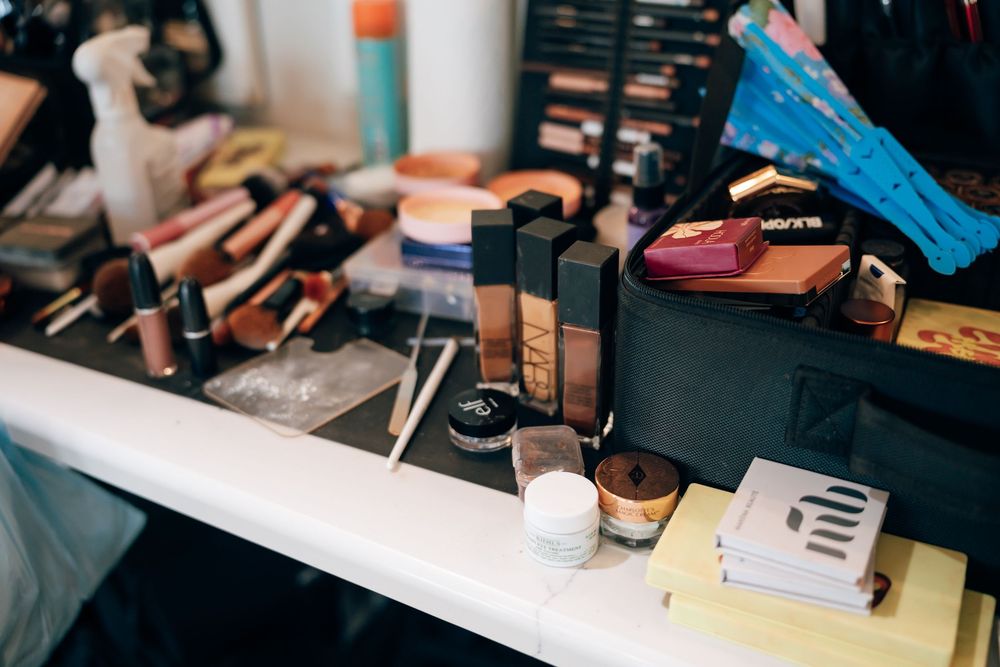 Makeup artist station with foundation bottles, brushes, palettes, and cosmetic products scattered across a white counter in a backstage stud
