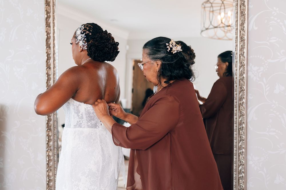 A woman in a brown cardigan helps a bride in a white lace gown with her dress in front of an ornate mirror.