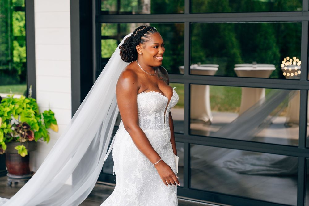 A smiling bride in a strapless white lace gown with a long veil stands near large windows with greenery in the background.