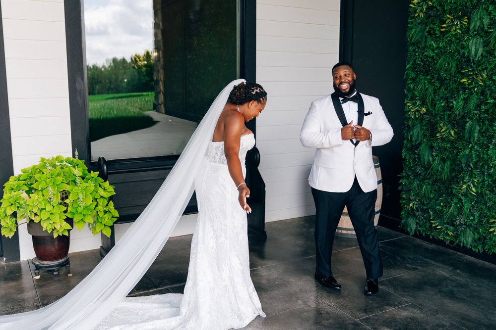 Bride in a lace gown with a long cathedral veil shares a joyful first look with a smiling groom in a white tuxedo jacket and black bow tie.