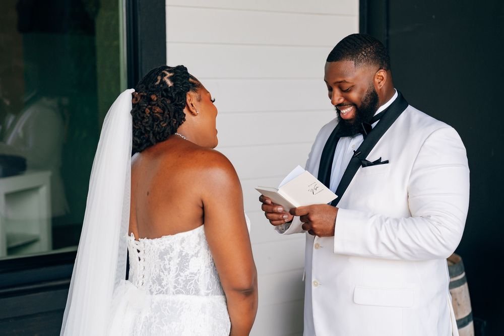 A groom in a white tuxedo jacket and black bow tie smiles while reading his vows to the bride in her wedding gown.
