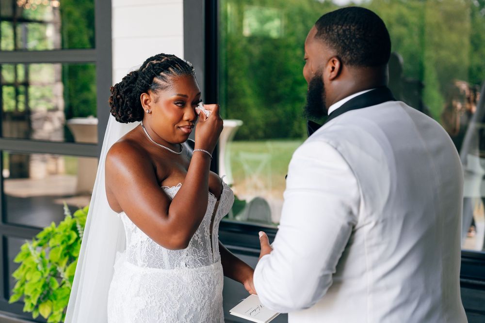 Emotional bride in a white lace wedding gown wipes a tear as she stands facing her groom in a white vest during their first look.